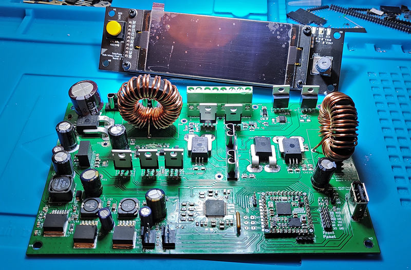 Photo of a circuit board with many components installed, including inductors and several ICs, with an LCD display propped up behind.